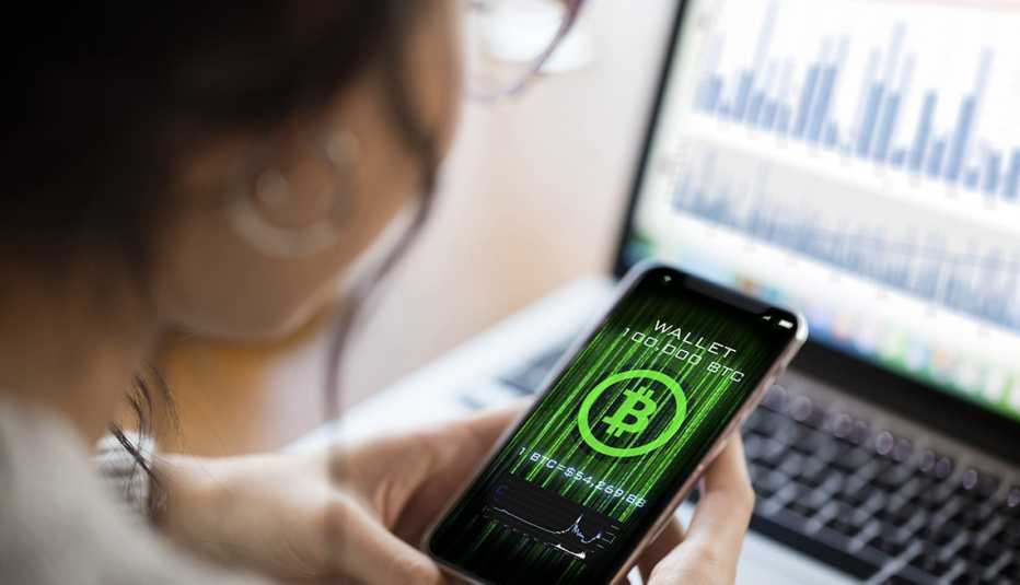 woman looking at mobile app display of a Bitcoin wallet in front of a laptop display of investment charts in soft focus 